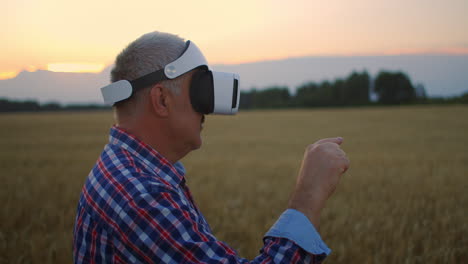An-elderly-male-farmer-uses-gestures-with-his-hands-standing-in-a-field-wearing-virtual-reality-glasses.-Use-VR-glasses-in-agriculture-on-a-field-with-wheat.